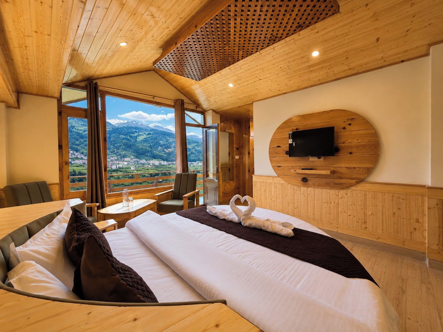 A hotel room with a large bed, a TV mounted on a wooden wall, and a view of a mountainous landscape.