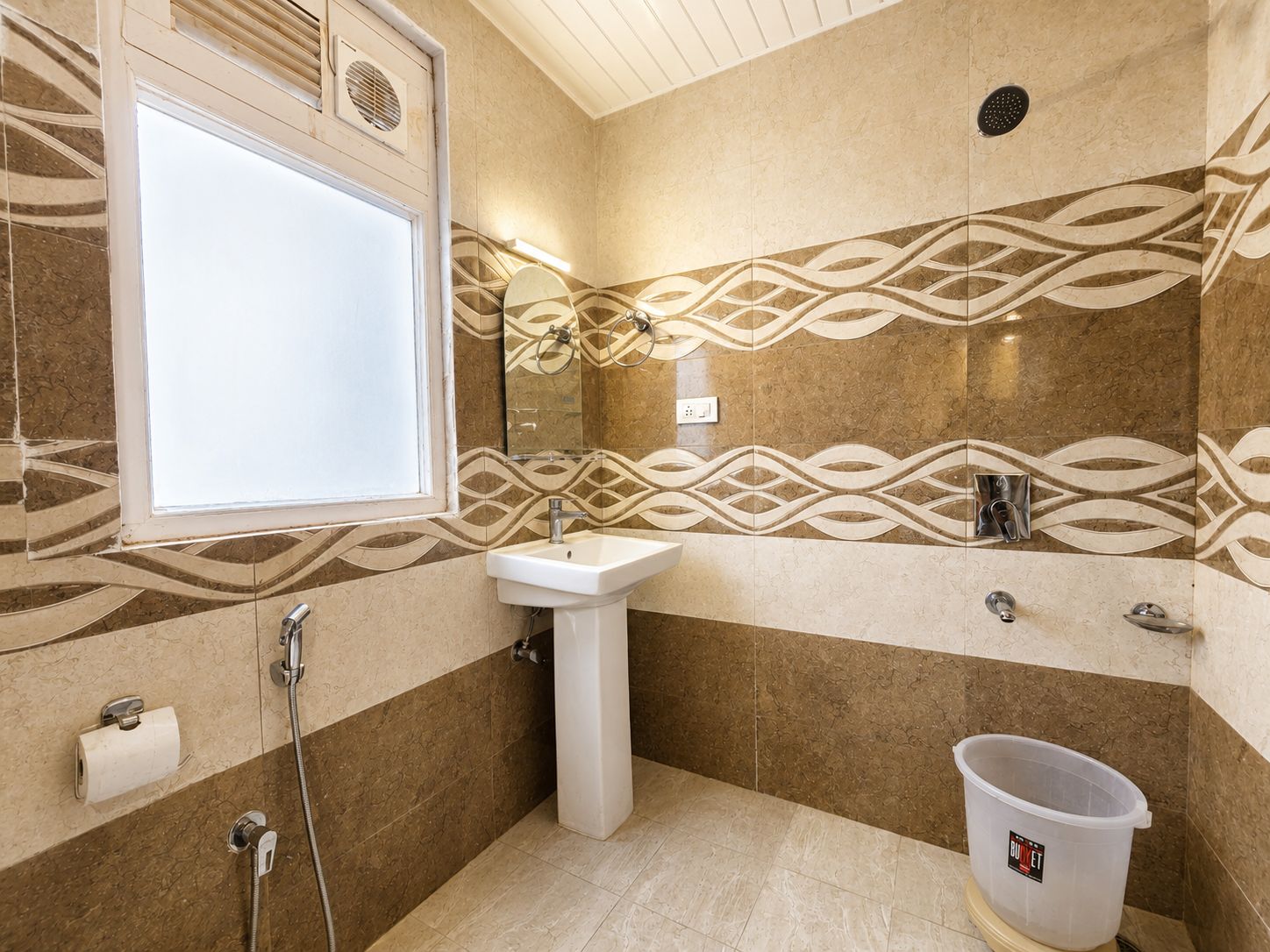 Et badeværelse med en hvid vask, bruser og toilet. Der er et vindue og et spejl over vasken.