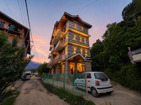 A multi-story building with balconies, a car parked in front, and a dirt road.