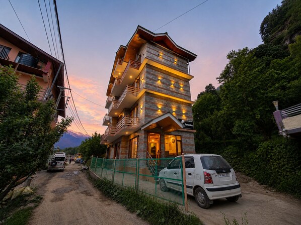 A multi-story building with balconies, a car parked in front, and a dirt road.