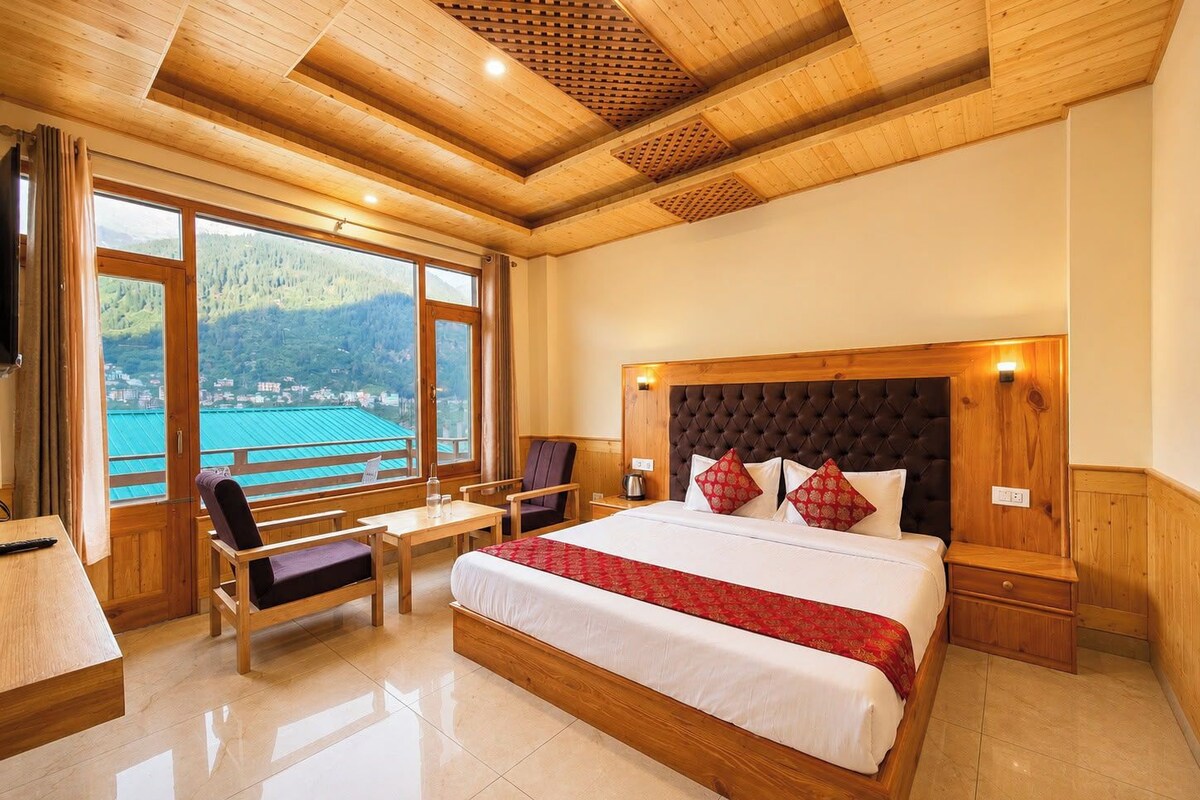 A spacious bedroom with a large bed, a wooden headboard, and a view of a mountainous landscape through large windows.