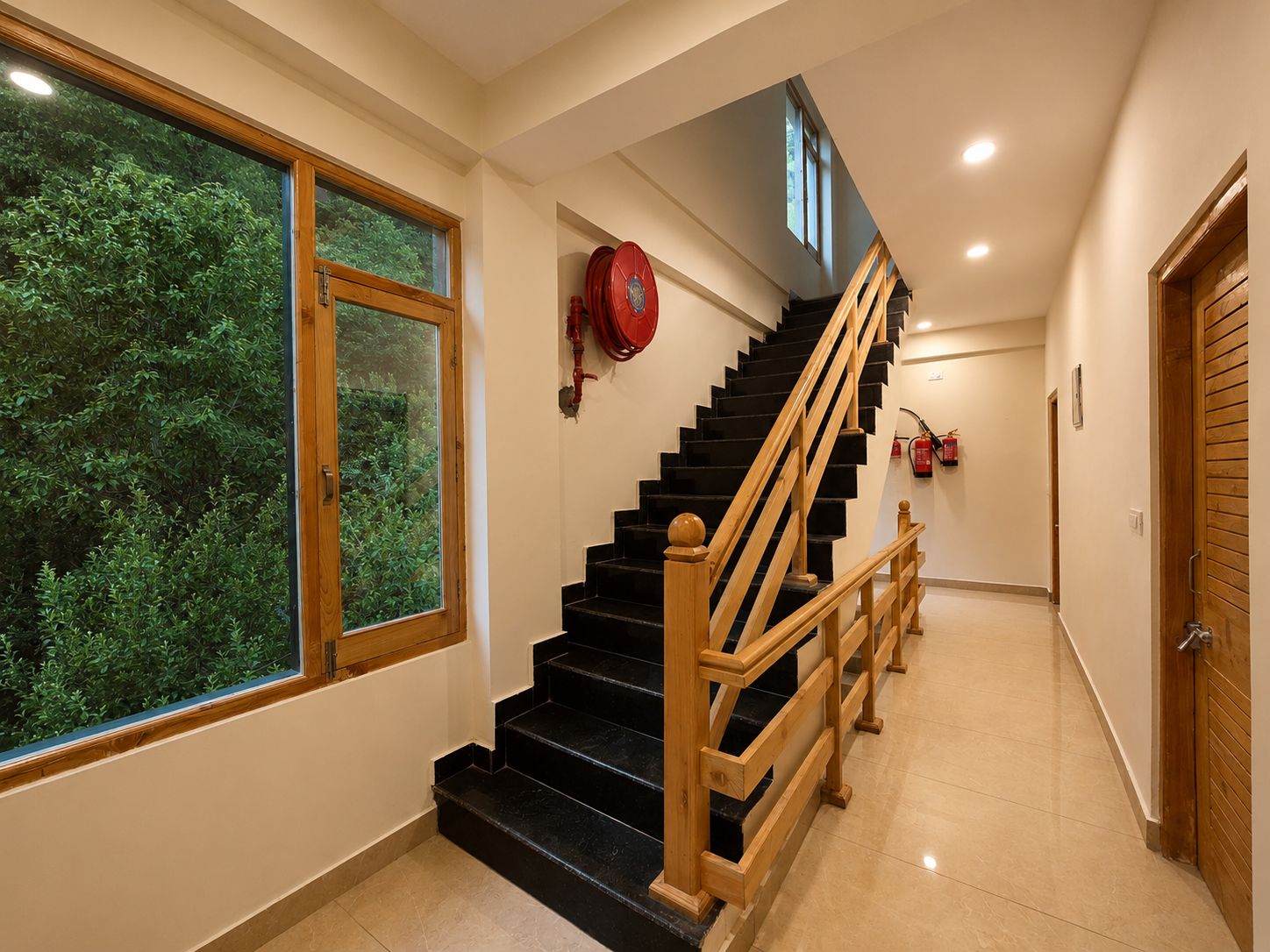 A staircase with wooden railings leading to an upper floor, a fire extinguisher mounted on the wall, and a window with a view of greenery.