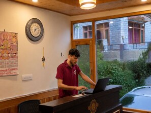 A person at a wooden desk with a laptop, a wall clock, and a poster in a room with a window and a door.