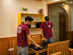 Two individuals in maroon shirts standing at a wooden table with a vase of flowers, in a room with wooden paneling and a yellow shelf with glassware.