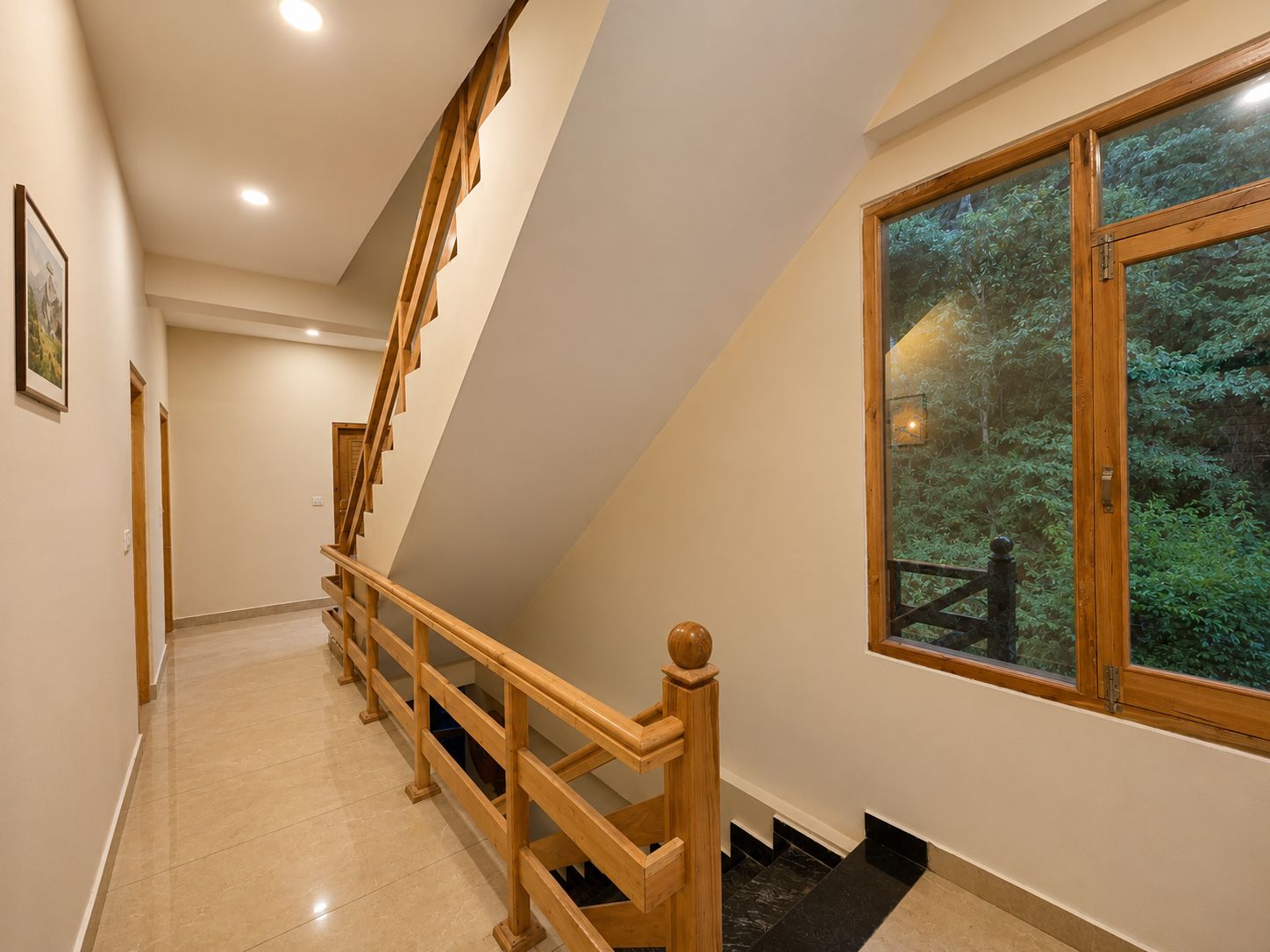 A staircase with wooden railings leading to a landing, a window with a view of greenery, and a framed picture on the wall.