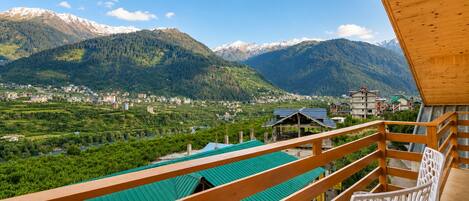 A balcony with a view of a mountainous landscape, a green-roofed building, and a white chair.