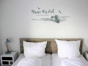 A bedroom with a bed, two pillows, a nightstand with a lamp, and a wall decal with text and a boat. - 50.4.s Waterkant (Wangerland)