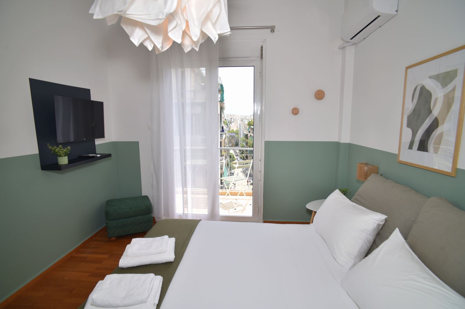 A hotel room with a bed, a TV mounted on the wall, a balcony with a view, and a painting on the wall.