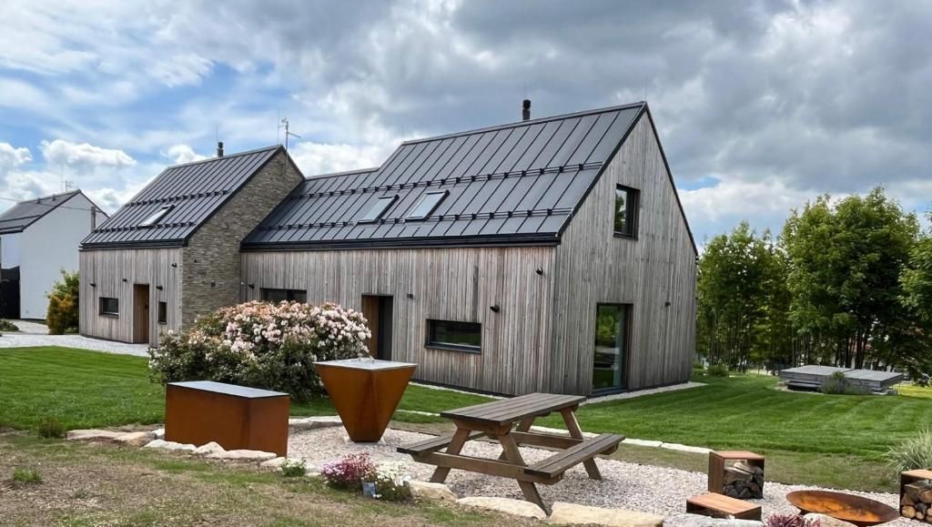 A modern house with a sloped roof, a large patio with a table and benches, and a garden with flowers and a fire pit.