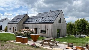 Una casa moderna con techo inclinado, un patio amplio con mesa y bancos, y un jardín con flores y un hogar para fuego.