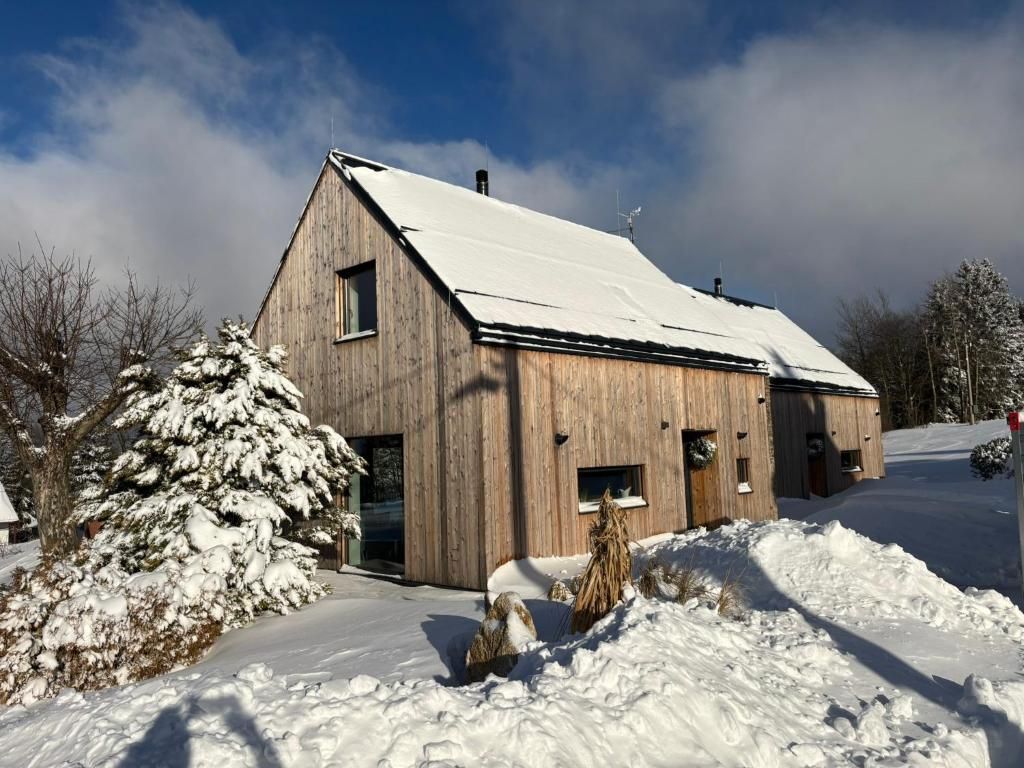 Une maison en bois avec un toit en pente recouvert de neige, entourée d’arbres enneigés et sous un ciel dégagé.