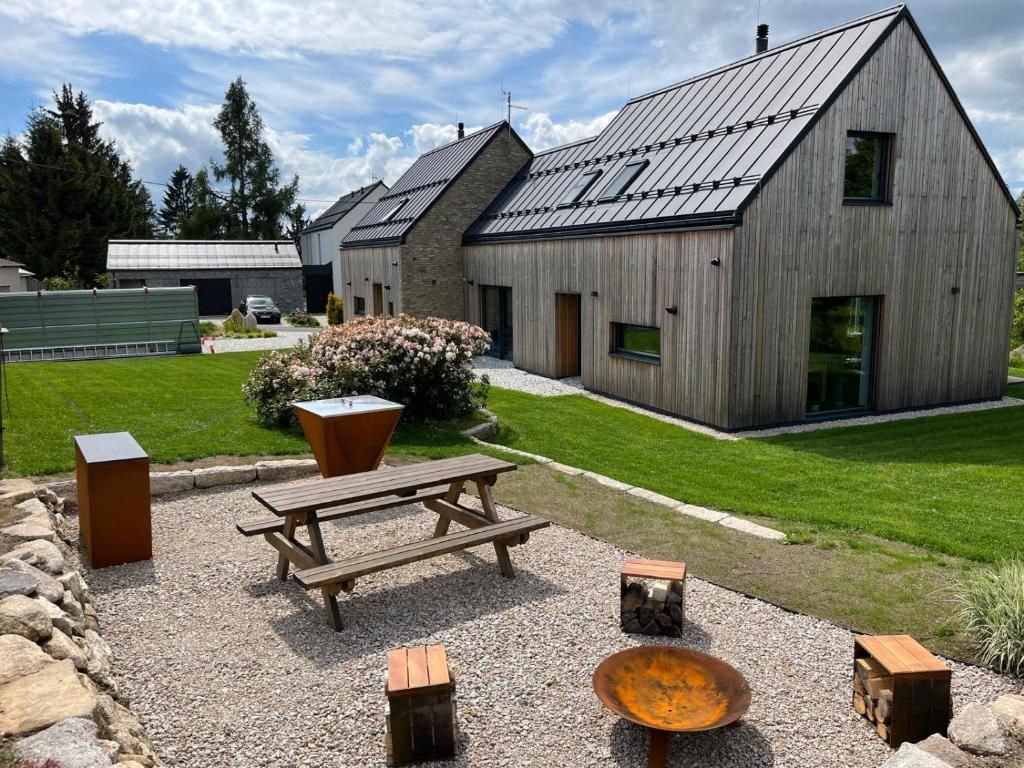 A modern building with a sloped roof, a gravel patio with a wooden table and benches, and a small fire pit.