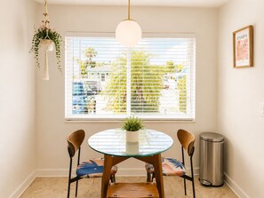 A dining area with a round glass table, two chairs, a hanging plant, a trash can, and a window with blinds. - Riviera Blue - The Palm Beaches Retreat - Florida, 2BR/2BTH (West Palm Beach)