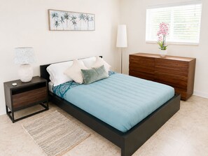 A bedroom with a bed, a nightstand, a dresser, a lamp, a window with blinds, and a painting on the wall. - Riviera Blue - The Palm Beaches Retreat - Florida, 2BR/2BTH (West Palm Beach)