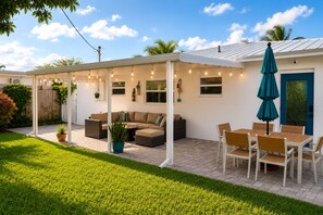 A covered patio area with a sectional sofa, dining table, and chairs, surrounded by potted plants and string lights. - Riviera Blue - The Palm Beaches Retreat - Florida, 2BR/2BTH (West Palm Beach)
