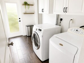A laundry room with a front-loading washer and dryer, a small trash bin, and a plant on a shelf.
