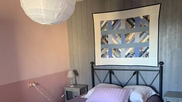 A bedroom with a bed, a nightstand, a lamp, a wall-mounted artwork, and a ceiling light fixture.