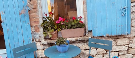 A stone building with a window, two blue chairs, a small table, and potted plants.