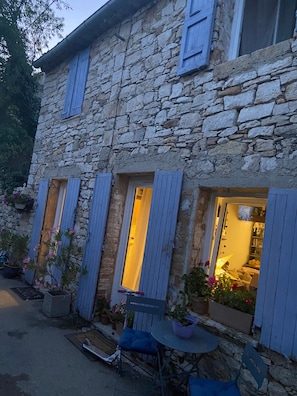 A stone building with multiple windows, some with shutters, and a small outdoor seating area with a table and chairs. - House 'La Petite Maison de Village' with Private Terrace and Wi-Fi in the Heart of Provence (Vachères)