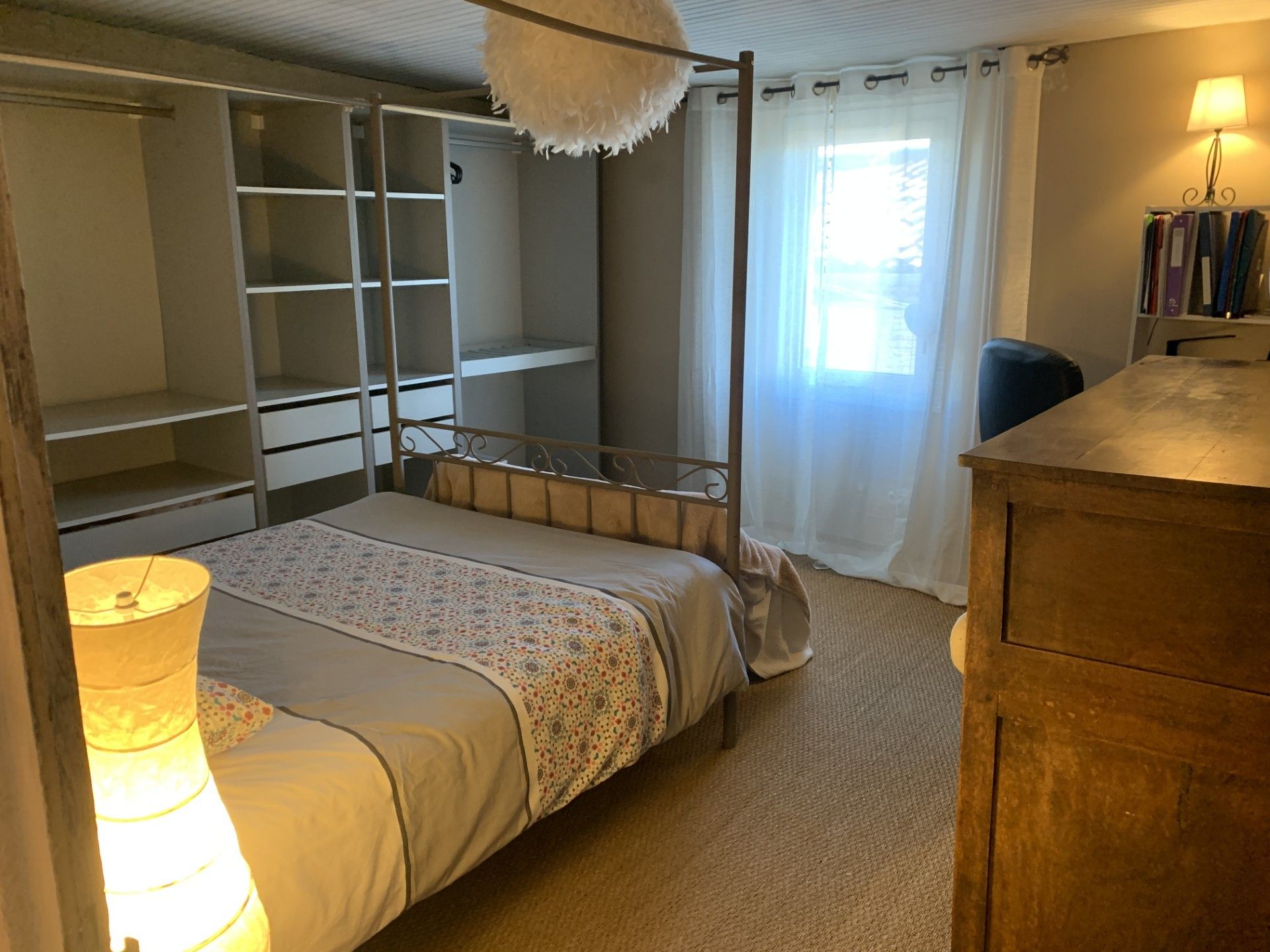 A bedroom with a four-poster bed, a wooden dresser, a bookshelf, and a window with curtains.