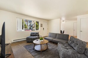 Una sala de estar moderna con sofá, otomanas y una mesa de centro, un televisor y un ventanal con vistas a edificios y árboles.