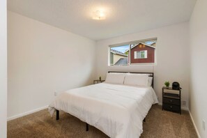 A bedroom with a single bed, a nightstand, a small table with a plant, and a window with a view of a red house. - Cozy 2BR Home in Lynnwood w  Backyard (Lynnwood)
