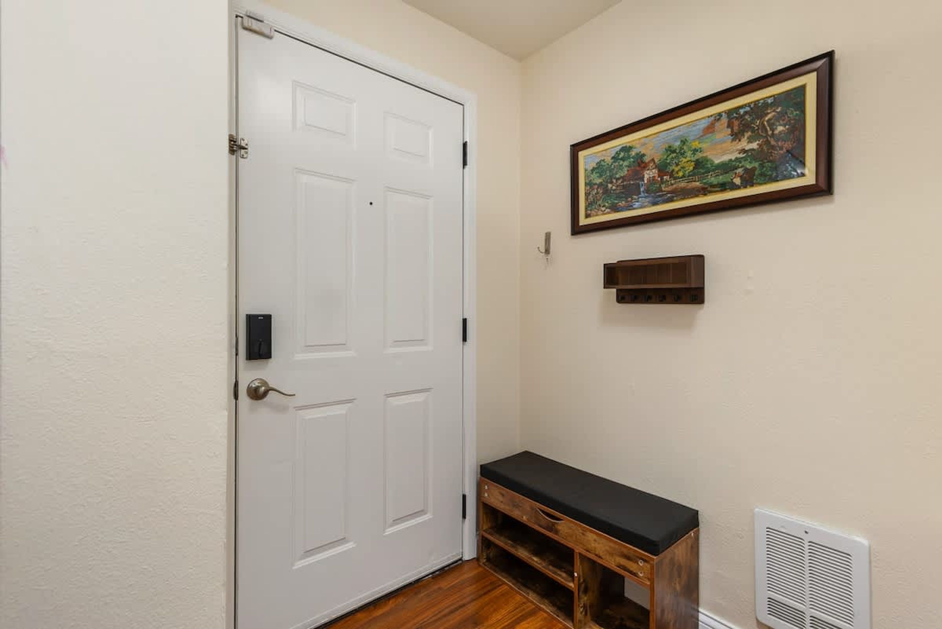 A hallway with a white door, a bench, a painting, and a vent.