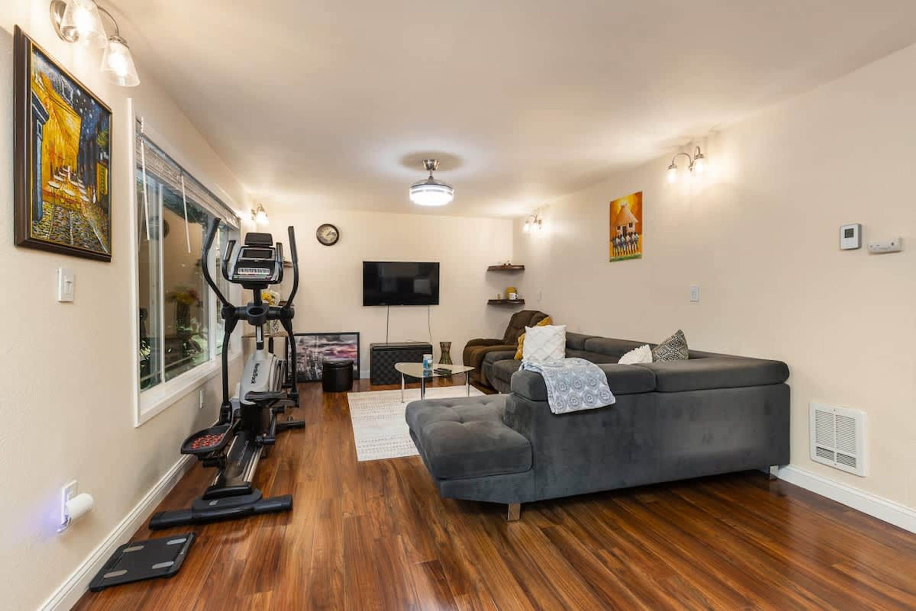 A living room with a grey sofa, a treadmill, and a flat-screen TV.