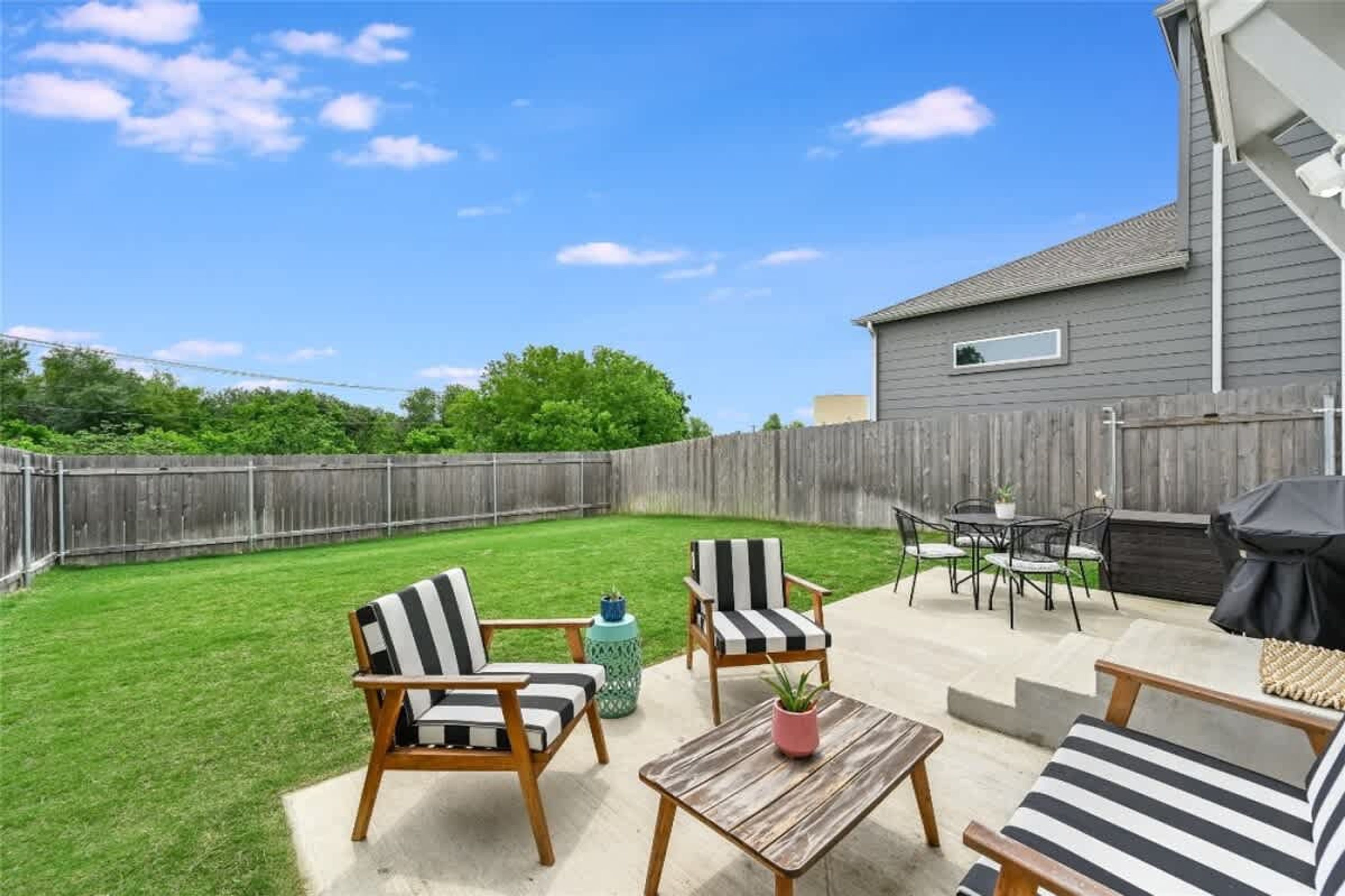 Een achtertuin met een houten tafel, twee gestreepte stoelen, een klein rond tafeltje en een barbecue.