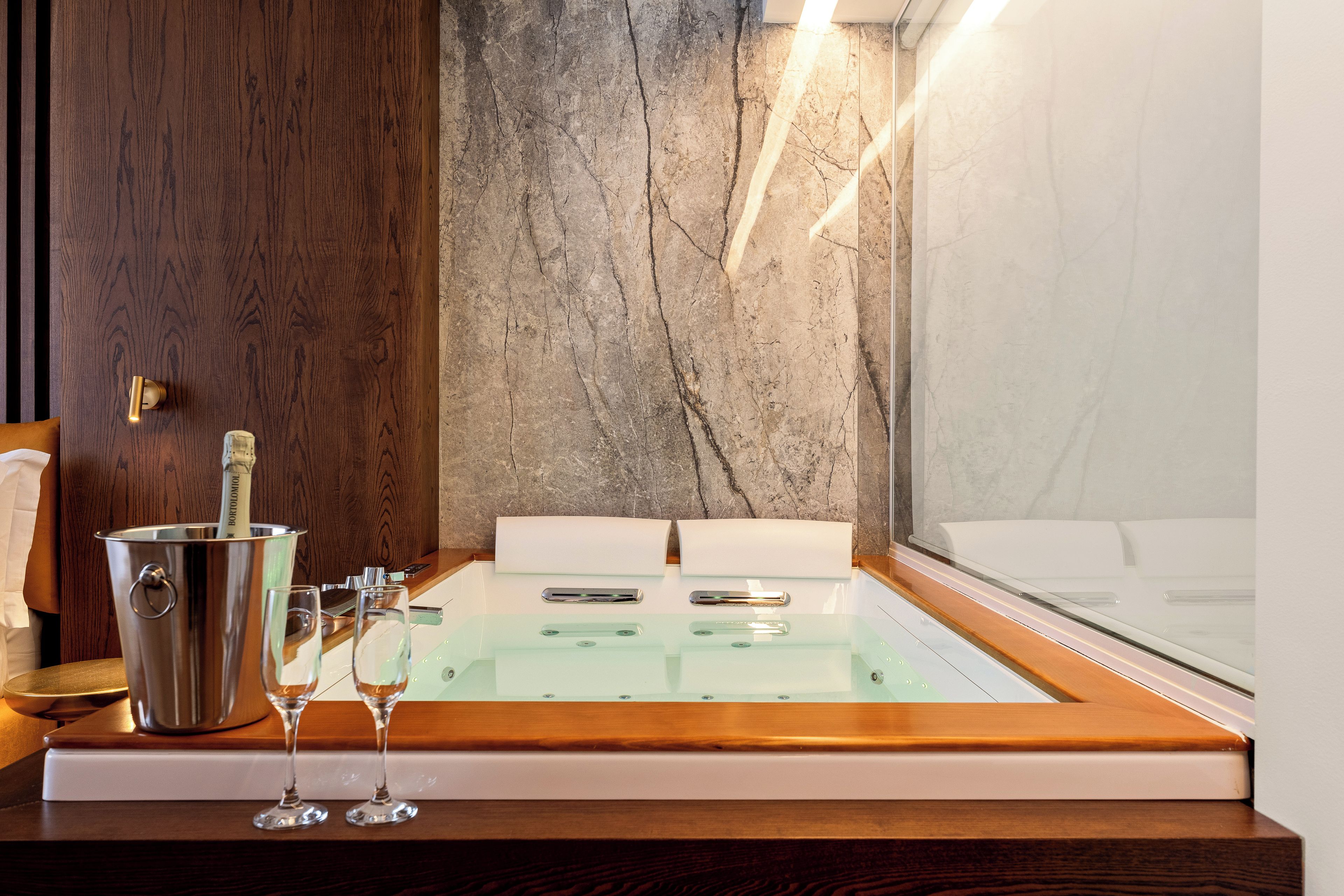 Un baño moderno con pared de mármol, suelo de madera, bañera de vidrio y cubo para champán.