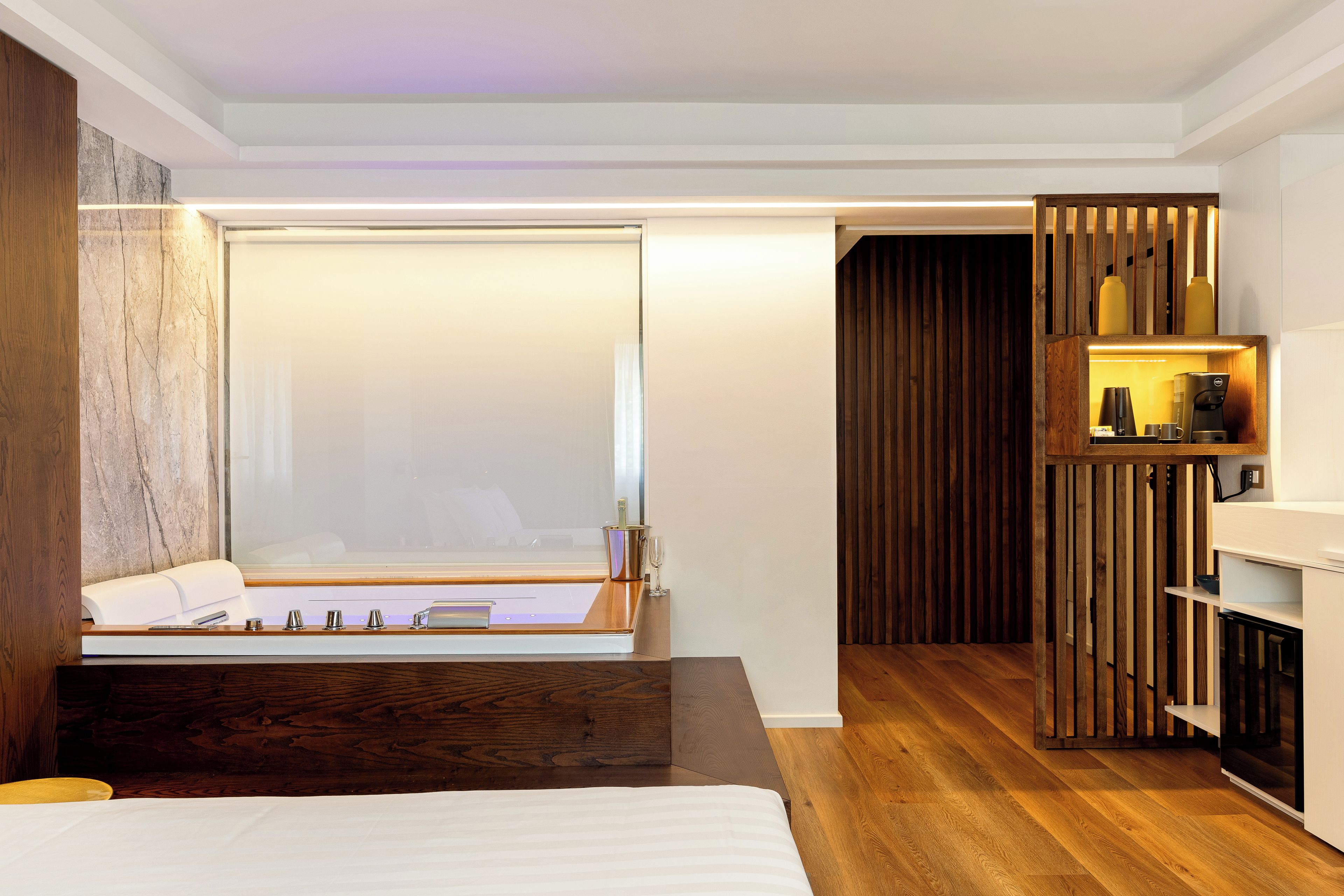 Ein modernes Hotelzimmer mit einem großen Bett, einem Nachttisch, einer Wand aus Holzpaneelen und einer integrierten Ablage mit einer Kaffeemaschine.