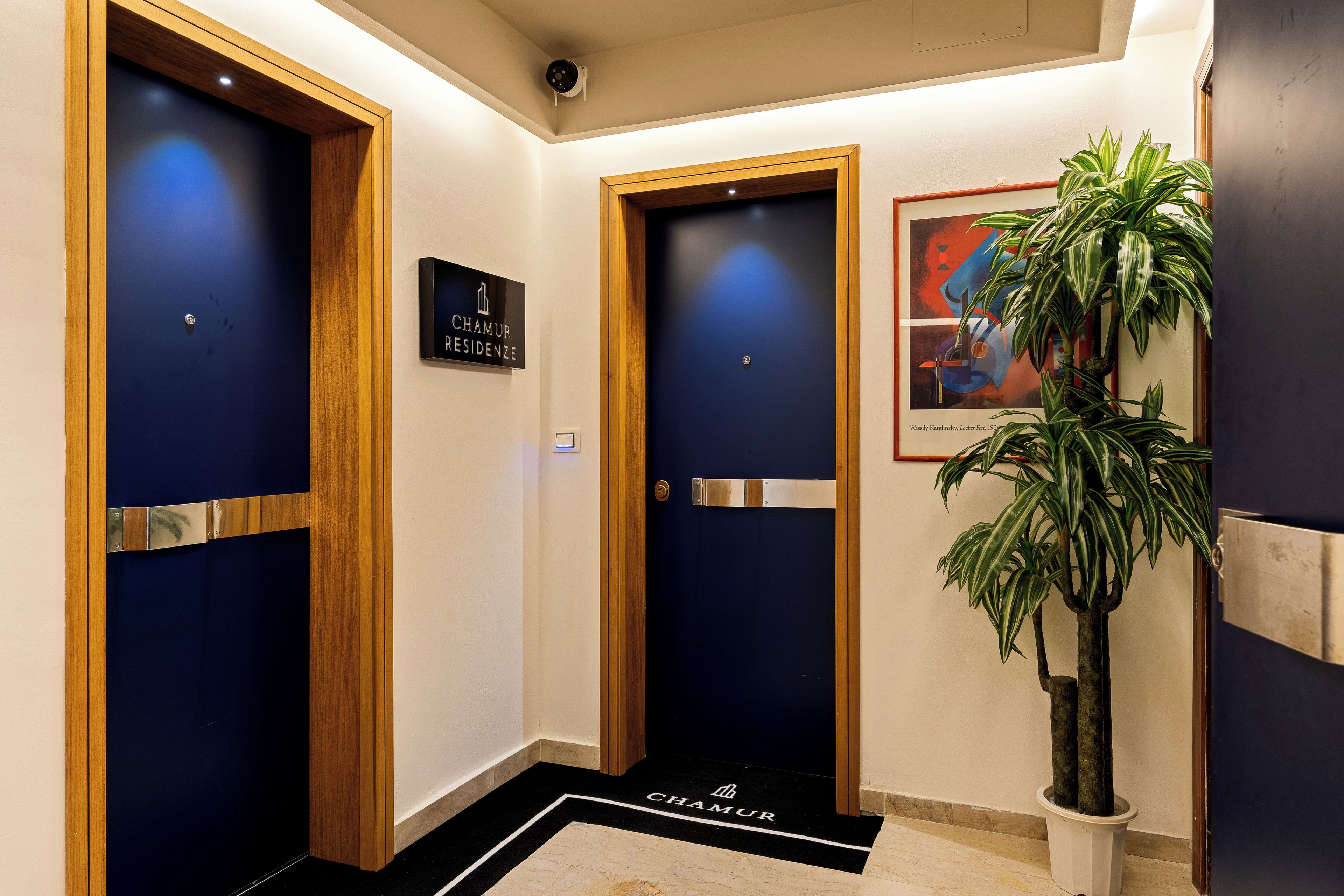 Un vestíbulo de hotel con dos puertas, un letrero sobre cada una, una planta en maceta y una obra de arte enmarcada en la pared.