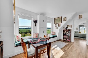 A dining area with a view of a green field, a wooden table with chairs, and a bookshelf with books and decorative items.