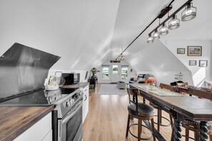 A modern kitchen with a large stove, a dining area with a wooden table and bar stools, and a loft bedroom with a skylight.