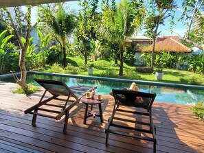 A wooden deck with two lounge chairs and a small table with two cups, overlooking a swimming pool surrounded by lush greenery and a house in the background. - An Bang Mystery - Art & Beach Villa (Đà Nẵng)