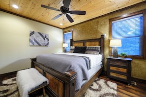 Ein Schlafzimmer mit Deckenventilator aus Holz, einem Bett mit grau-weiß gestreifter Bettdecke, einem kleinen Nachttisch mit Lampe und einem Fenster mit Jalousien.