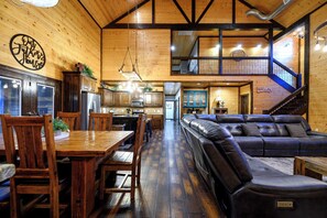 A spacious living area with a large leather sofa, wooden dining table, and chairs. A kitchen with modern appliances is visible in the background. - 4 King Bedrooms | Hot Tub | Game Room (Broken Bow)