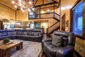 A spacious living area with a large L-shaped sectional sofa, a smaller sofa, and a coffee table. A staircase leads to an upper level with a balcony. - 4 King Bedrooms | Hot Tub | Game Room (Broken Bow)