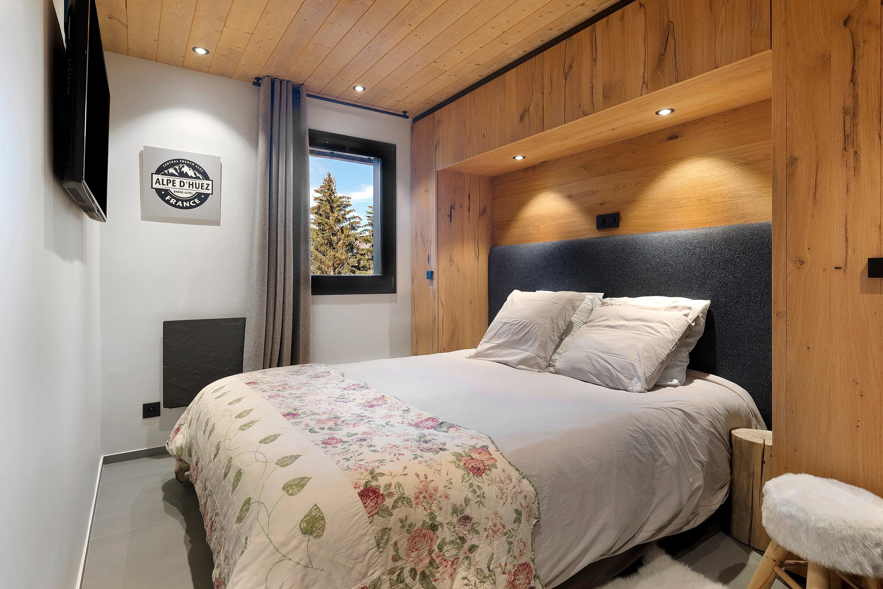 Un dormitorio con cama, ventana con vista a árboles, televisor montado y techo de madera.