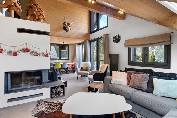 A modern interior with a fireplace, a sofa, a dining area, and large windows offering views of the outdoors.