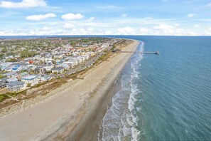 An aerial view of a coastal area with a long pier extending into the sea, adjacent to a densely populated beachfront with multi-story buildings. - Ocean-View North Beach Condo with Pools & Community Private Beach Access (Tybee Island)