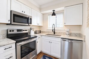 A modern kitchen with white cabinetry, stainless steel appliances, a granite countertop, and a window with blinds. - Ocean-View North Beach Condo with Pools & Community Private Beach Access (Tybee Island)