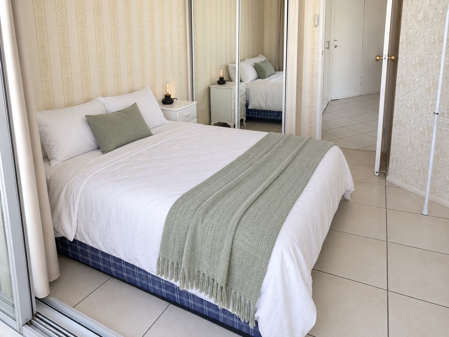A neatly made bed with a white and green blanket, a bedside table with a lamp, and a mirror reflecting the room.