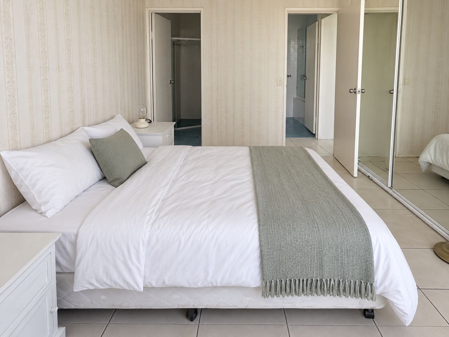 A neatly made bed with white linens and a grey throw, a bedside table with a lamp, and a large mirror on the wall.