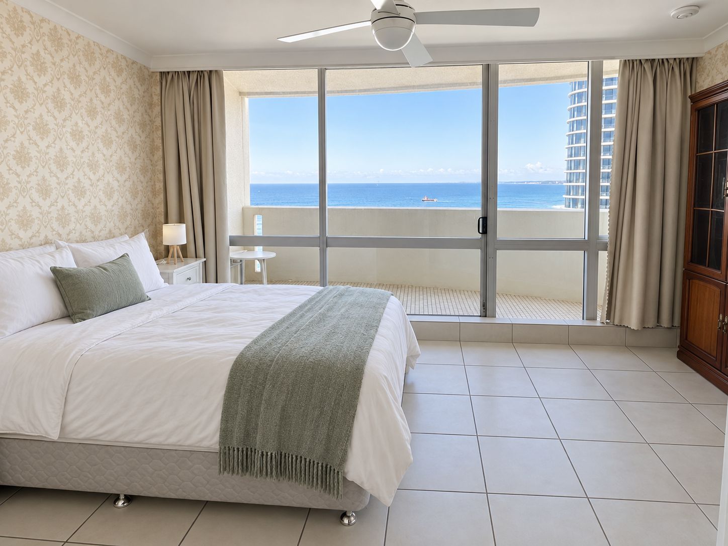 Ein Hotelzimmer mit Bett, Nachttisch, Lampe, Meerblick und einem Balkon mit Vorhängen.
