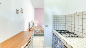 A room with a wooden dining table, a bed with a patterned bedspread, and a kitchenette with a gas stove. - Apartment 'Le Colonie - Pineta' with Private Terrace and Shared Garden (Presicce)