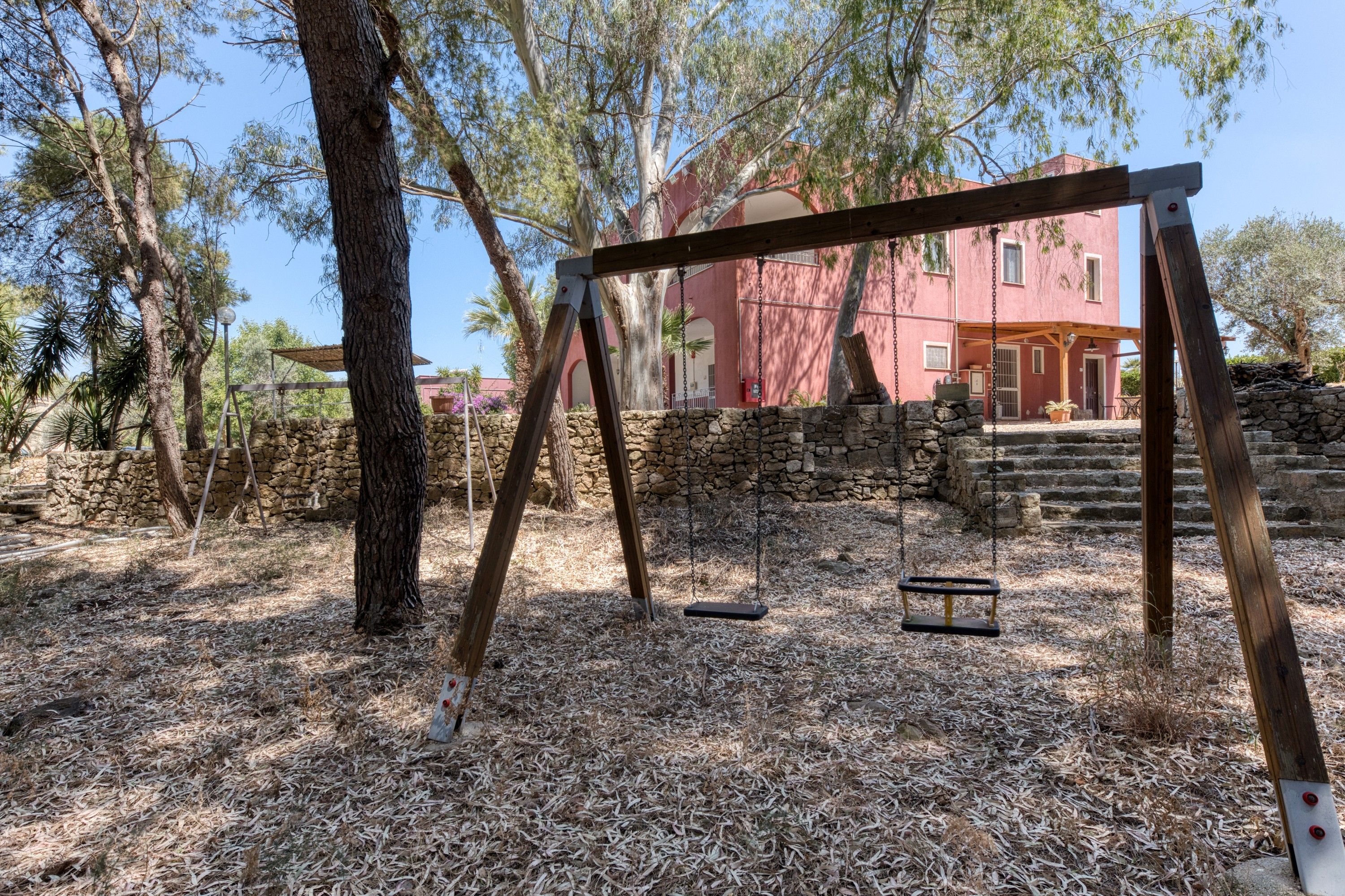 Un ensemble de balançoires en bois, installé dans un espace boisé, avec un mur de pierres et un bâtiment rouge en arrière-plan.