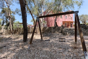 Un ensemble de balançoires en bois, installé dans un espace boisé, avec un mur de pierres et un bâtiment rouge en arrière-plan.