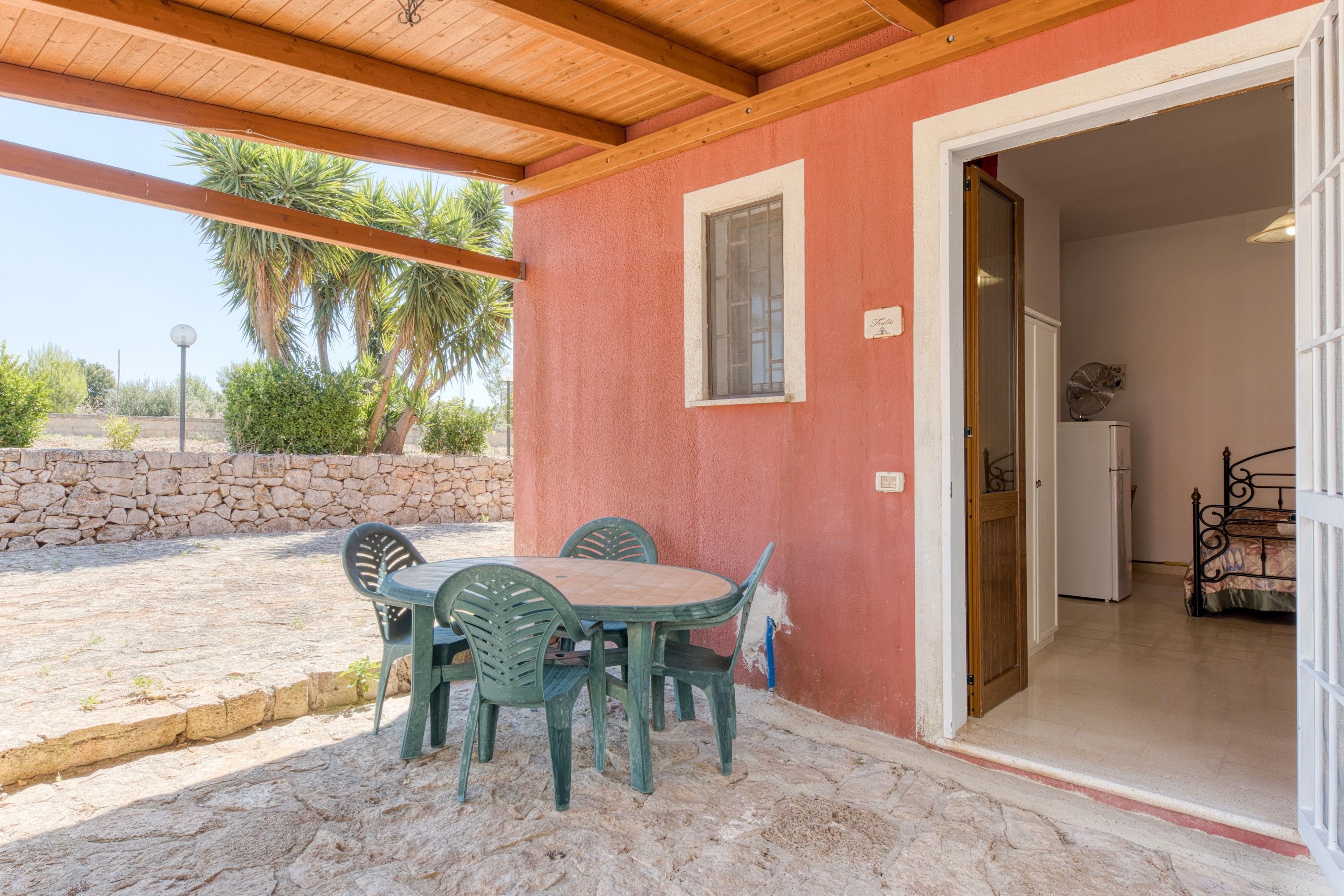 Un'area patio coperta con tavolo e sedie, un muro in pietra e una porta che dà su una camera da letto con un letto.