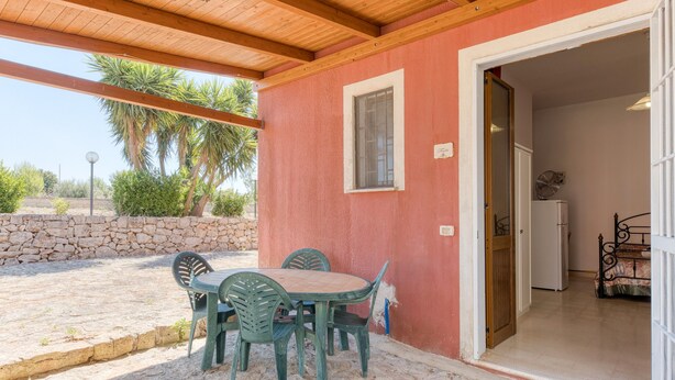 A covered patio area with a table and chairs, a stone wall, and a door leading to a bedroom with a bed. - Apartment 'Le Colonie - Pineta' with Private Terrace and Shared Garden (Presicce)